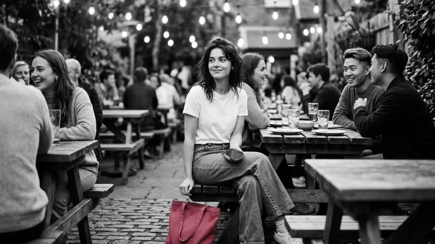 En un vespre càlid, al jardí d’un pub ple de gent, una jove d’uns vint-i-dos anys amb una samarreta blanca senzilla i uns texans amples seu atapeïda en un banc entre amics de la seva edat que riuen, amb el mòbil a la mà, pantalla avall, i un somriure tranquil i esperançat mentre aparta un moment la mirada de la conversa. A terra, a prop dels seus peus, hi ha una bossa de lona senzilla de color rosa intens, a sobre pengen garlandes de llums, i no hi ha cap rètol ni lletres a la imatge.